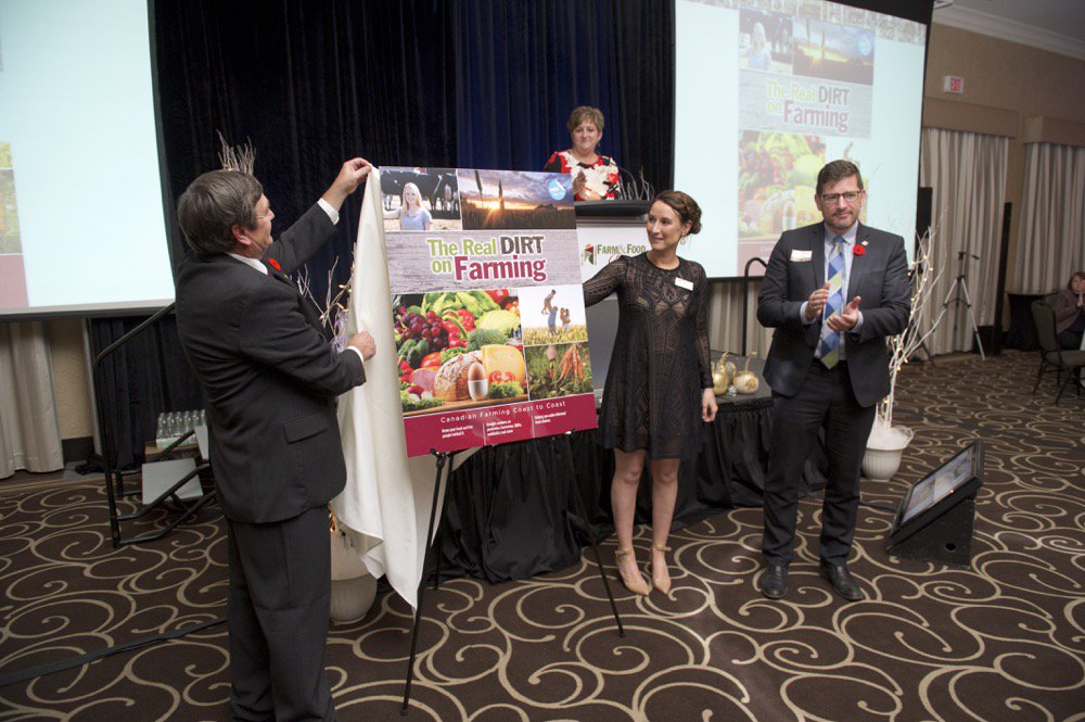 The new cover of the Real Dirt on Farming was unveiled at the Farm & Food Care Harvest Gala last week. Doing the unveiling was Farm & Food Care chair Brian Gilroy, left, Christine Schoonderwoerd, vice chair, and representing funder Wallenstein Feed & Supply Ltd., Pierre Petelle, president of CropLife Canada. Farm & Food Care Executive Director Kelly Daynard is at the podium.  Photo courtesy Farm and Food Care.
