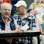 A man sitting at a table grins widely after winning a hard and fast bidding war.