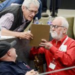 A gentleman leans over a table while a judge, in a bright red shirt with silver hair and beard, shares the components of how competitors are judged.