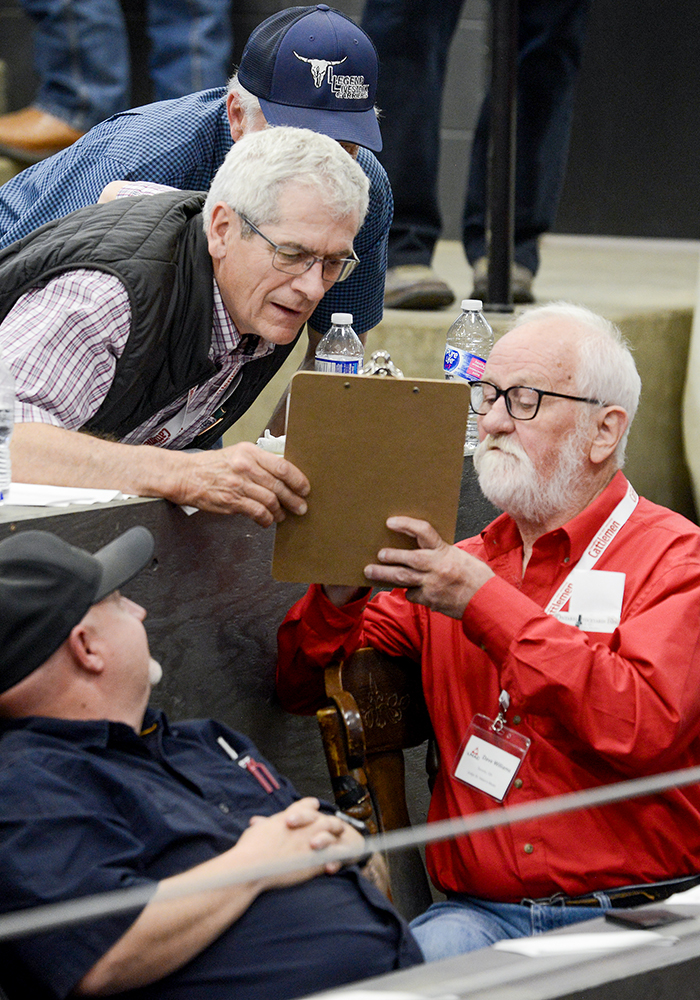 A gentleman leans over a table while a judge, in a bright red shirt with silver hair and beard, shares the components of how competitors are judged.