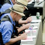 A row of three men in blue shirts, black vests and straw fedoras log what price each draft of cattle sells for.