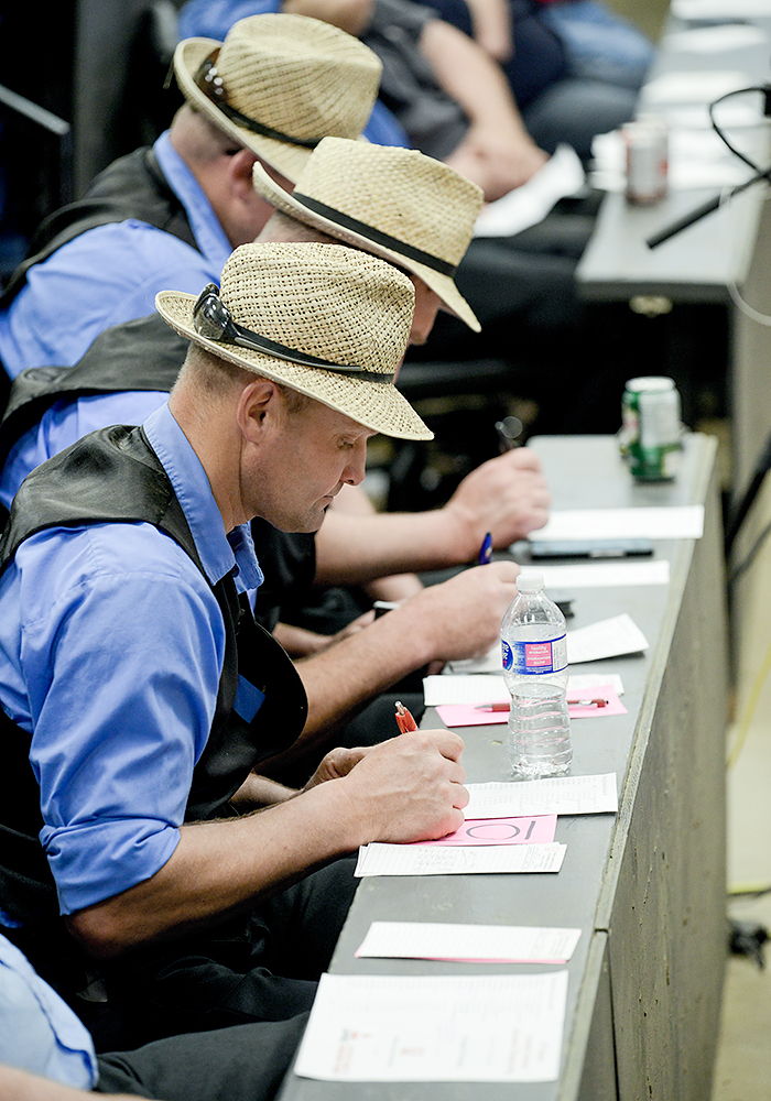 A row of three men in blue shirts, black vests and straw fedoras log what price each draft of cattle sells for.