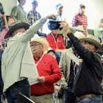 A man in a light suit jacket and cowboy hat raises his arm to select a piece of paper from a ball cap held up by a man in a dark suit jacket and a cowboy hat.