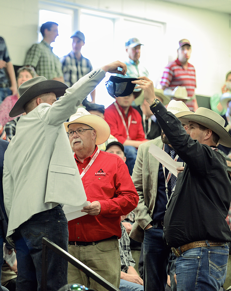 A man in a light suit jacket and cowboy hat raises his arm to select a piece of paper from a ball cap held up by a man in a dark suit jacket and a cowboy hat.