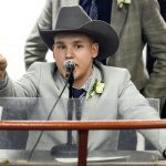 A man in a light suit jacket and charcoal coloured cowboy hat chants into the microphone while pointing out bids