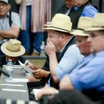 a man in a blue shirt, black vest and straw fedora covers his mouth as he yawns