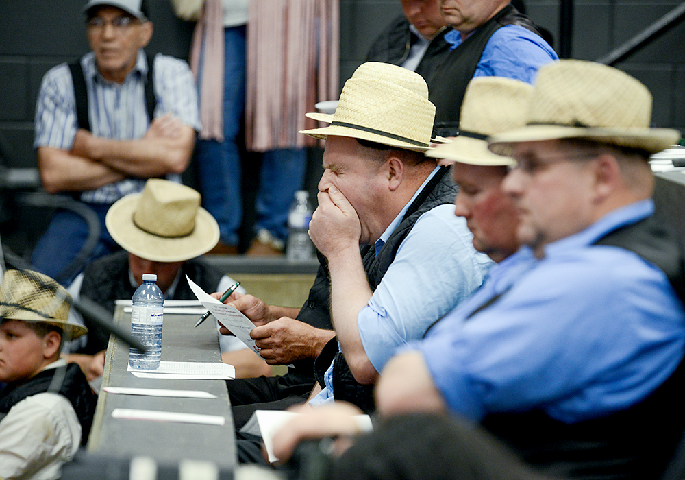 a man in a blue shirt, black vest and straw fedora covers his mouth as he yawns