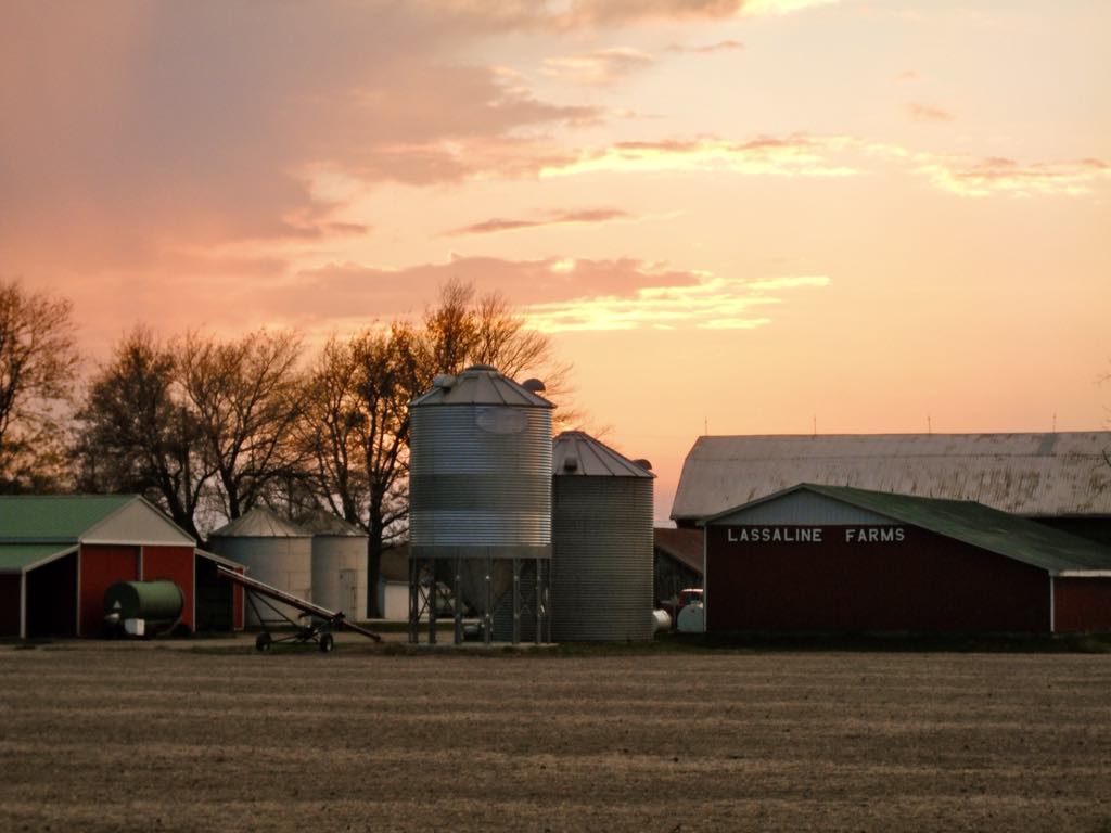 A picturesque farm featuring a red barn and silos, illuminated by the warm hues of sunset.